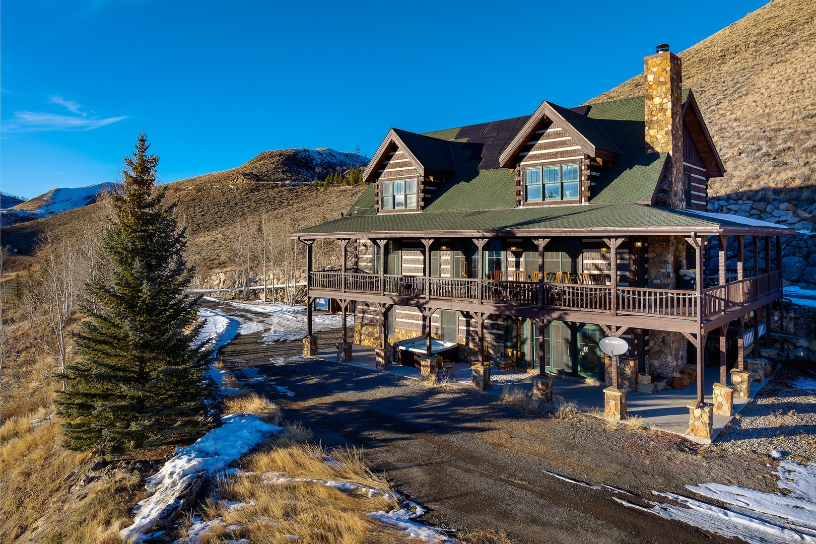Mountain lodge with expansive wraparound porches and stone chimney, nestled in rolling hills with winter snow patches.