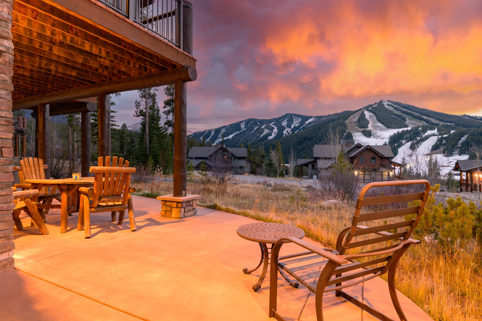 Mountain lodge exterior with patio seating area featuring dramatic sunset sky over snow-covered ski slopes and surrounding forest.