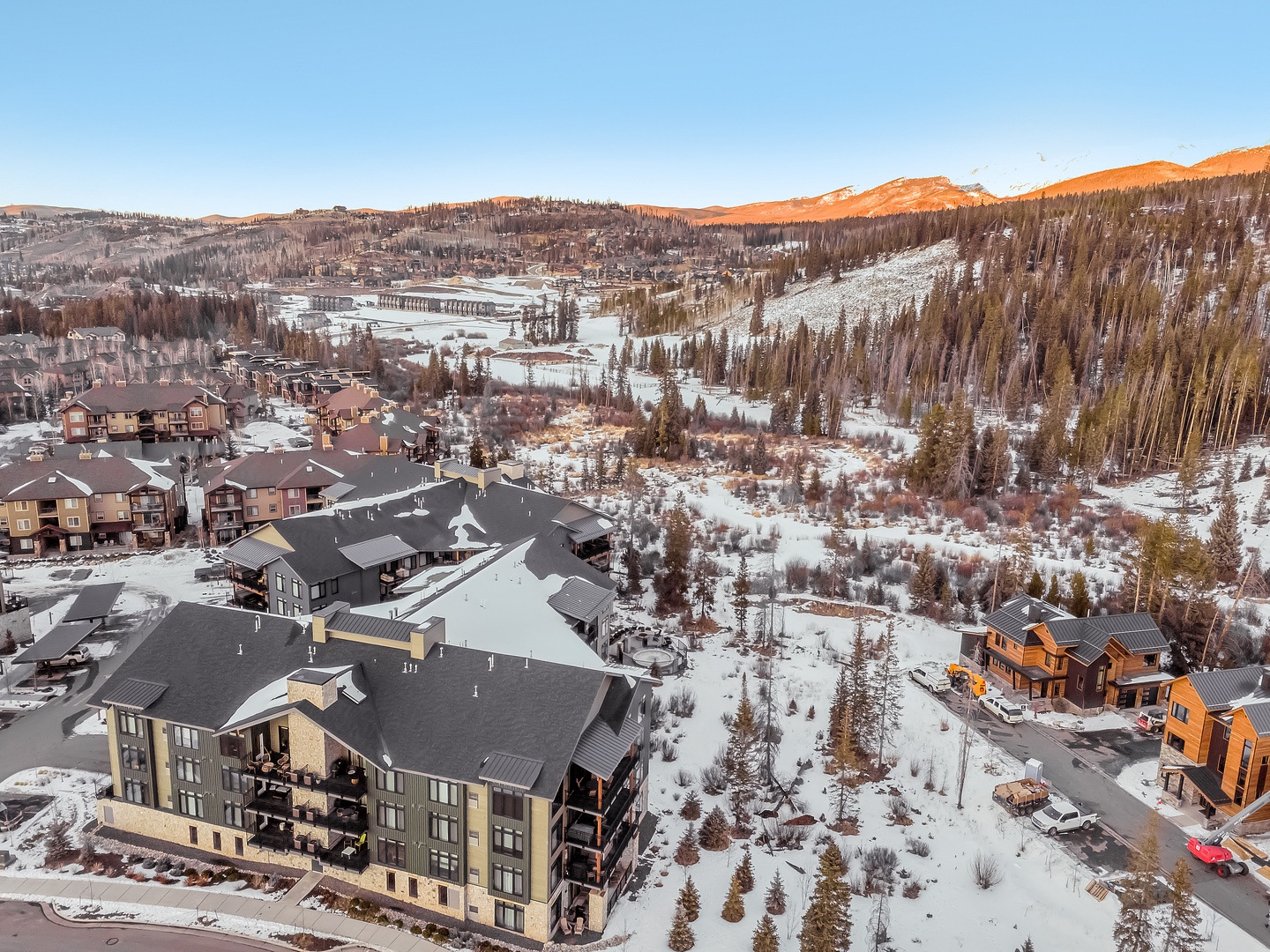 Aerial view showcases the property nestled in a snow-covered mountain valley with modern residential buildings and forested hills.