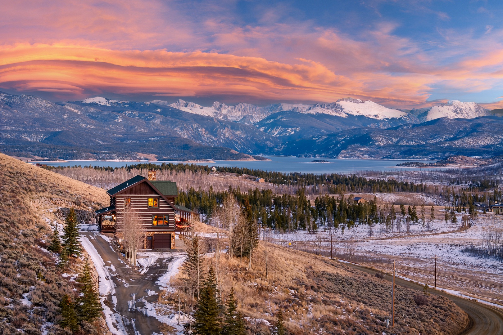 Mountain lodge nestled in winter landscape with snow-capped peaks and vibrant sunset sky over pristine wilderness valley.