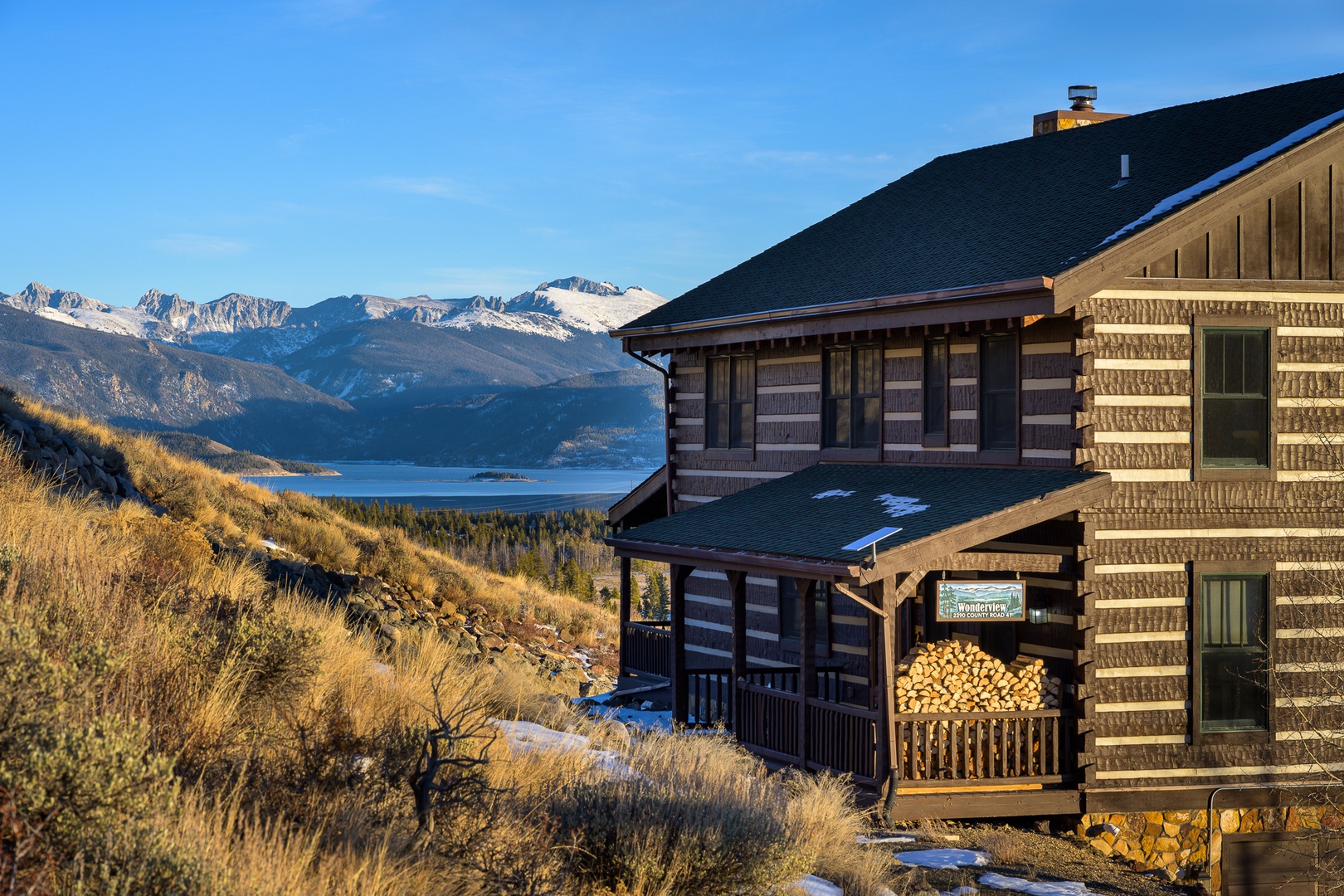 Rustic log cabin overlooking snow-capped peaks and pristine mountain lake with golden evening light.