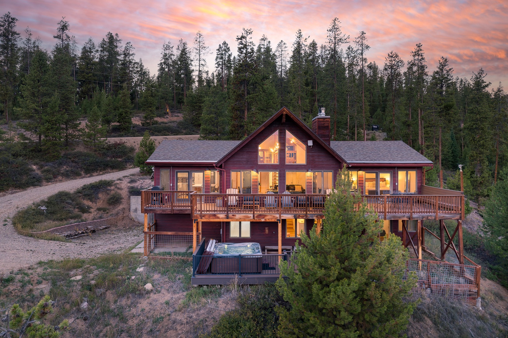 A welcoming mountain cabin illuminated against a stunning sunset sky, featuring expansive wraparound decks and a private hot tub nestled among towering pines in a peaceful wooded setting.