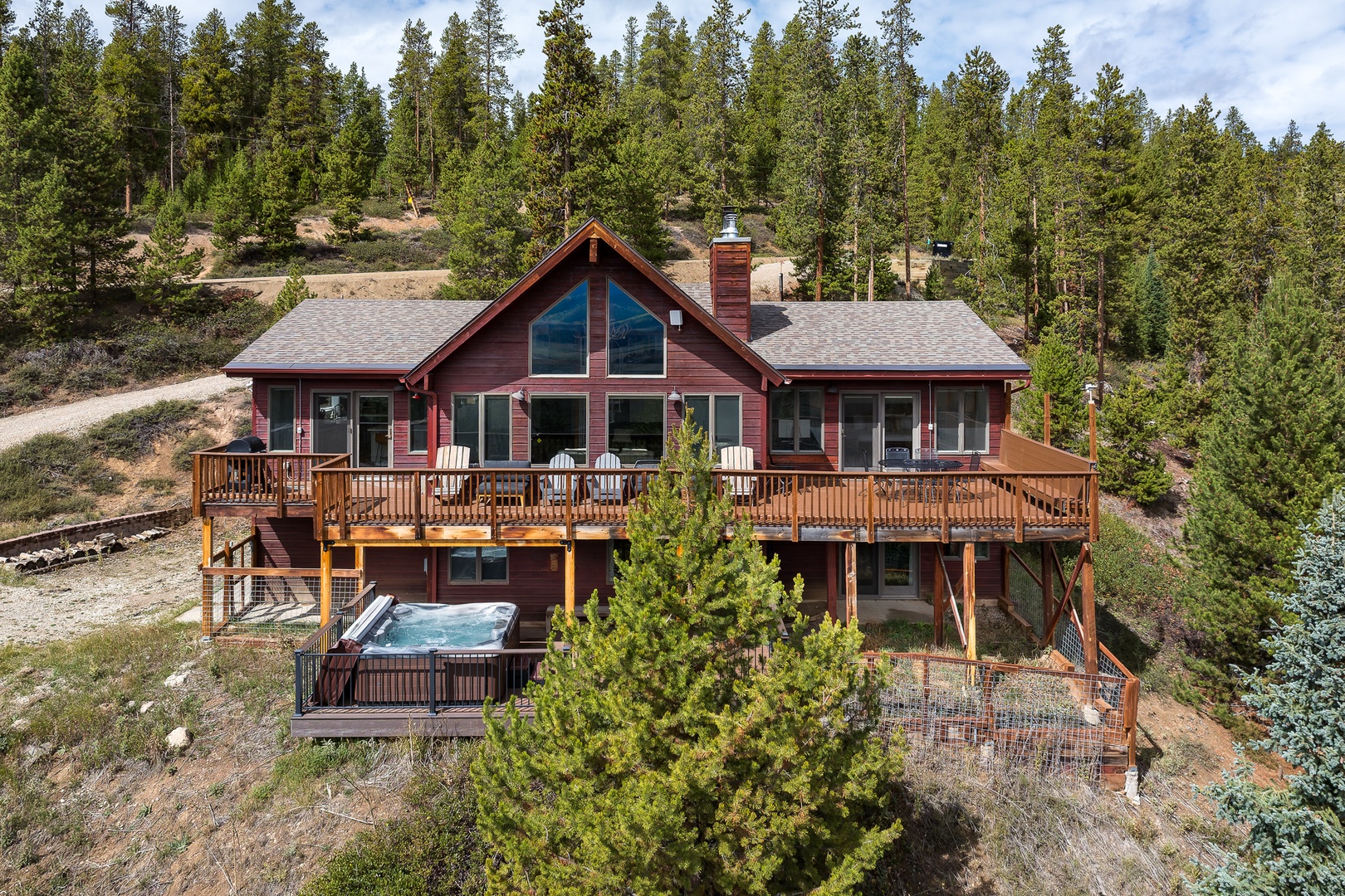 An elevated aerial view showcasing the complete property exterior nestled in a forested hillside setting, featuring expansive wooden decking, multiple levels, and an outdoor hot tub surrounded by mature evergreen trees and natural landscape.