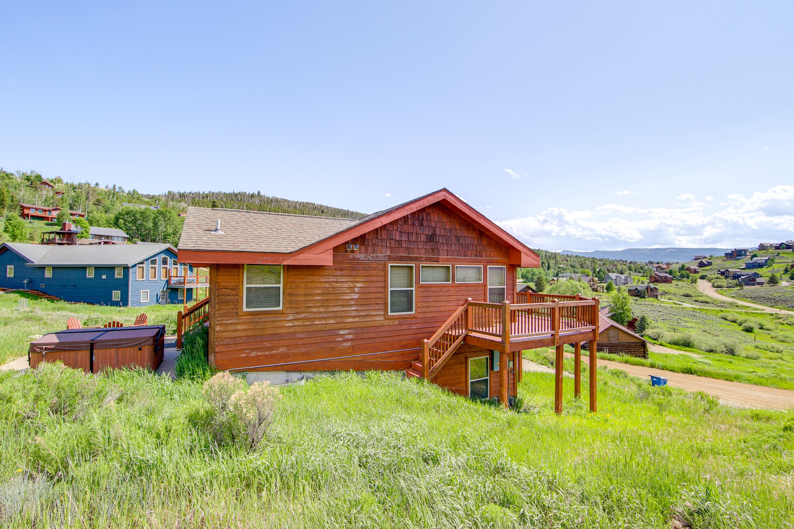 Mountain retreat with wooden siding and wraparound deck nestled among rolling hills and neighboring homes in a scenic valley setting.