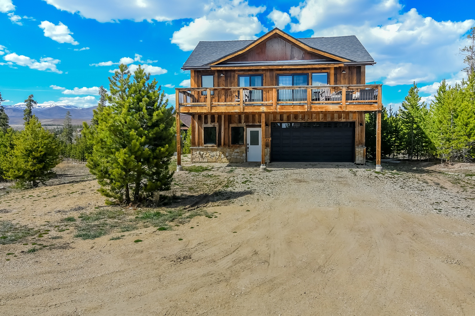 Modern mountain retreat featuring rustic wood construction, spacious deck, and garage, surrounded by evergreen trees with mountain backdrop.