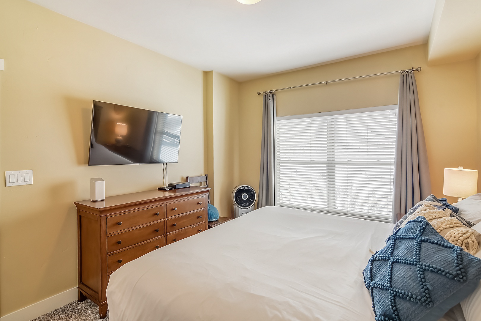 Drift off in your cozy bedroom featuring a plush bed, warm linens, flat-screen TV, and gentle natural light filtering through blinds.