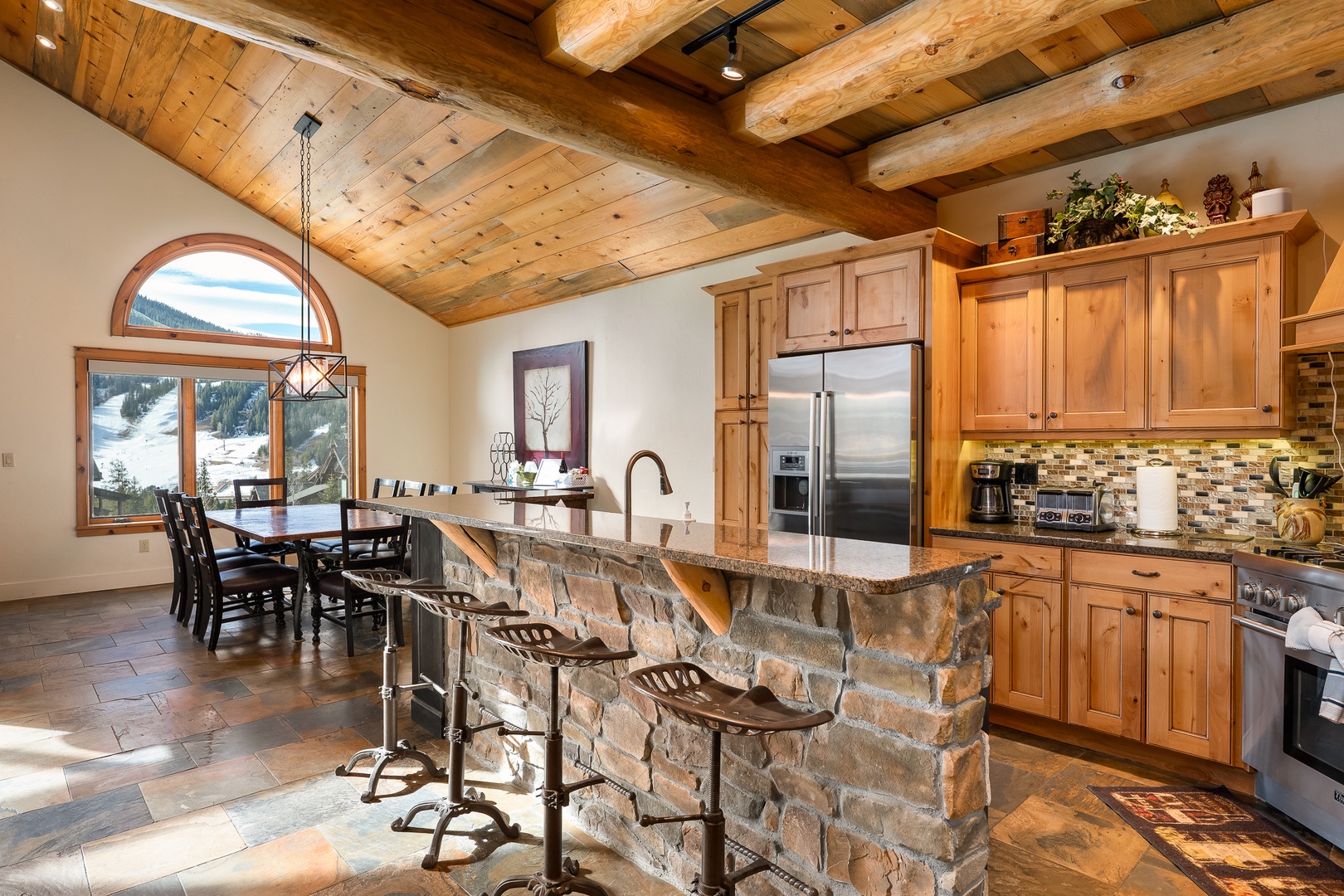 Gather in your stunning mountain kitchen with exposed wood beams, stone island, and panoramic ski slope views through arched windows.