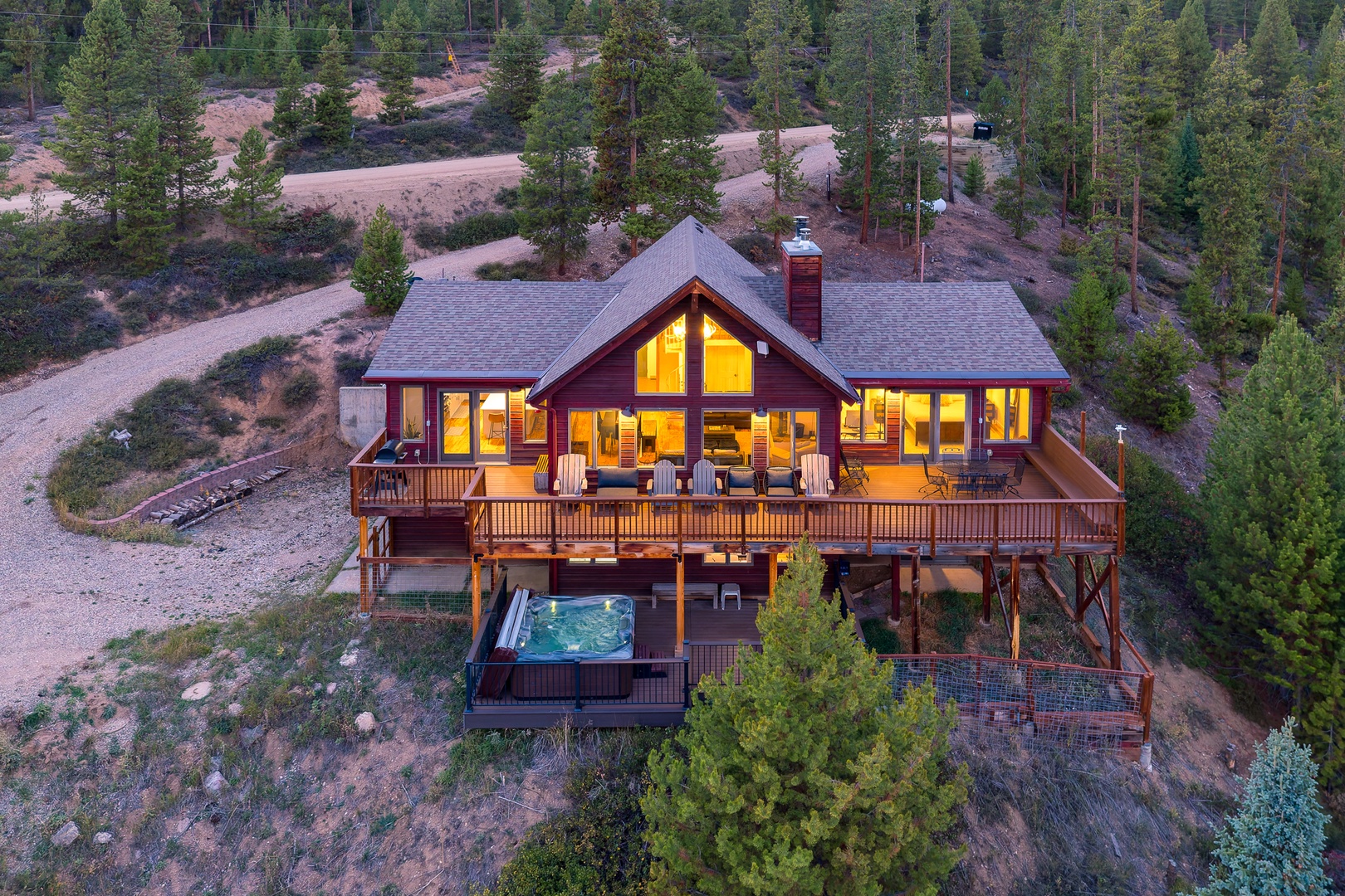 A striking mountain retreat with expansive deck and hot tub, nestled among tall pines at the edge of serene wilderness. Warm interior lighting welcomes as natural surroundings embrace this peaceful escape.