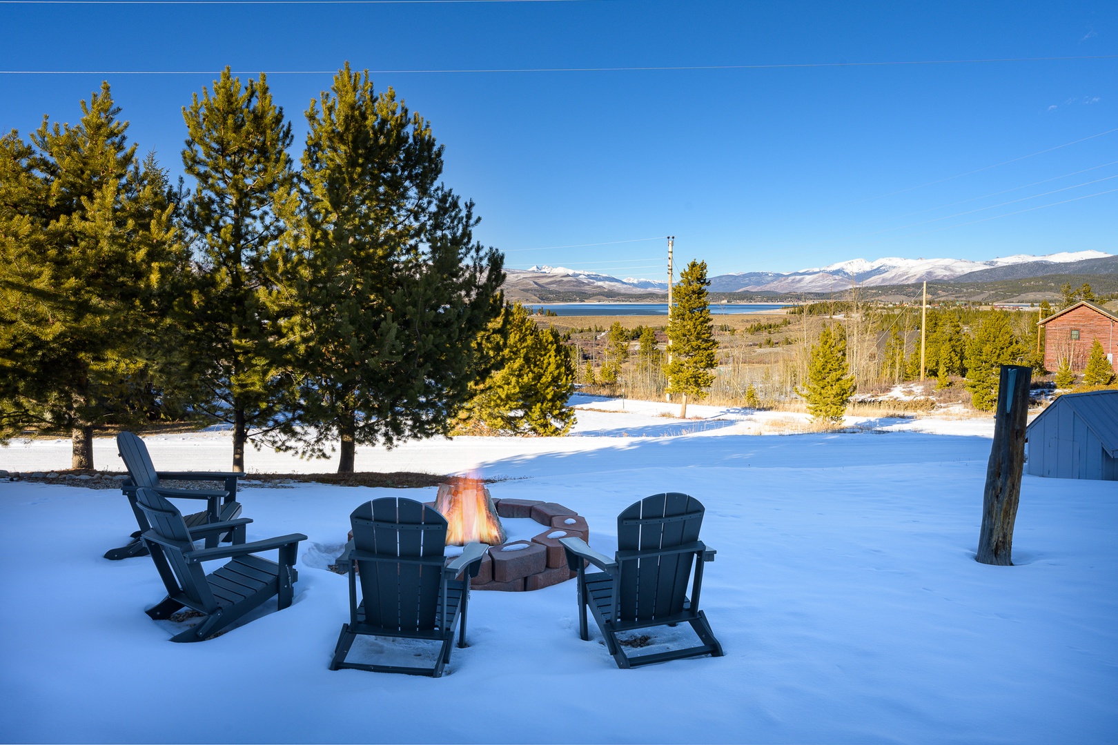 Snowy winter landscape features a fire pit surrounded by Adirondack chairs, with mountain views and evergreen trees creating a scenic mountain retreat setting.