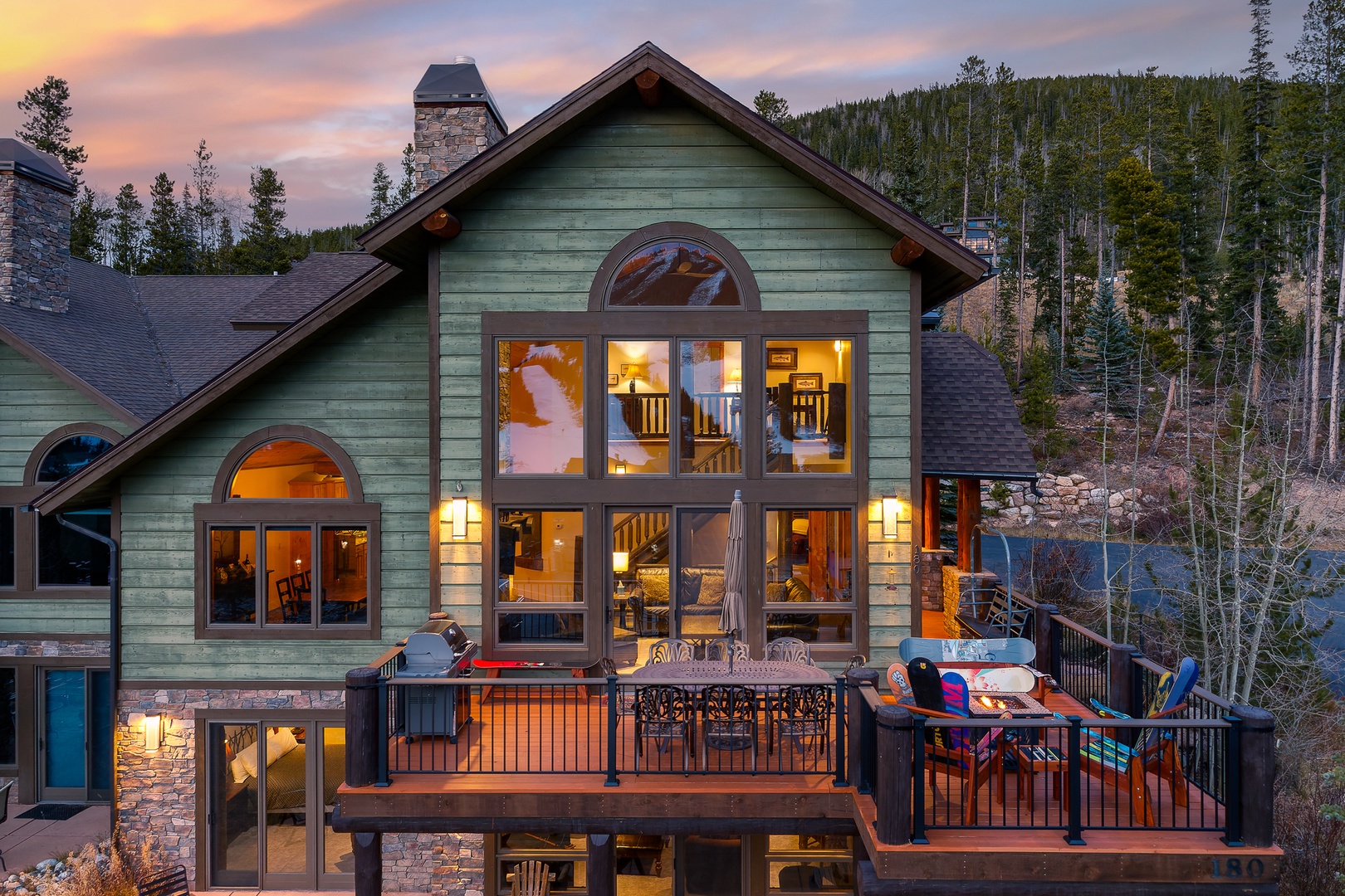 Mountain retreat glowing warmly at sunset, nestled among towering pines with expansive windows and elevated deck overlooking the forested valley.