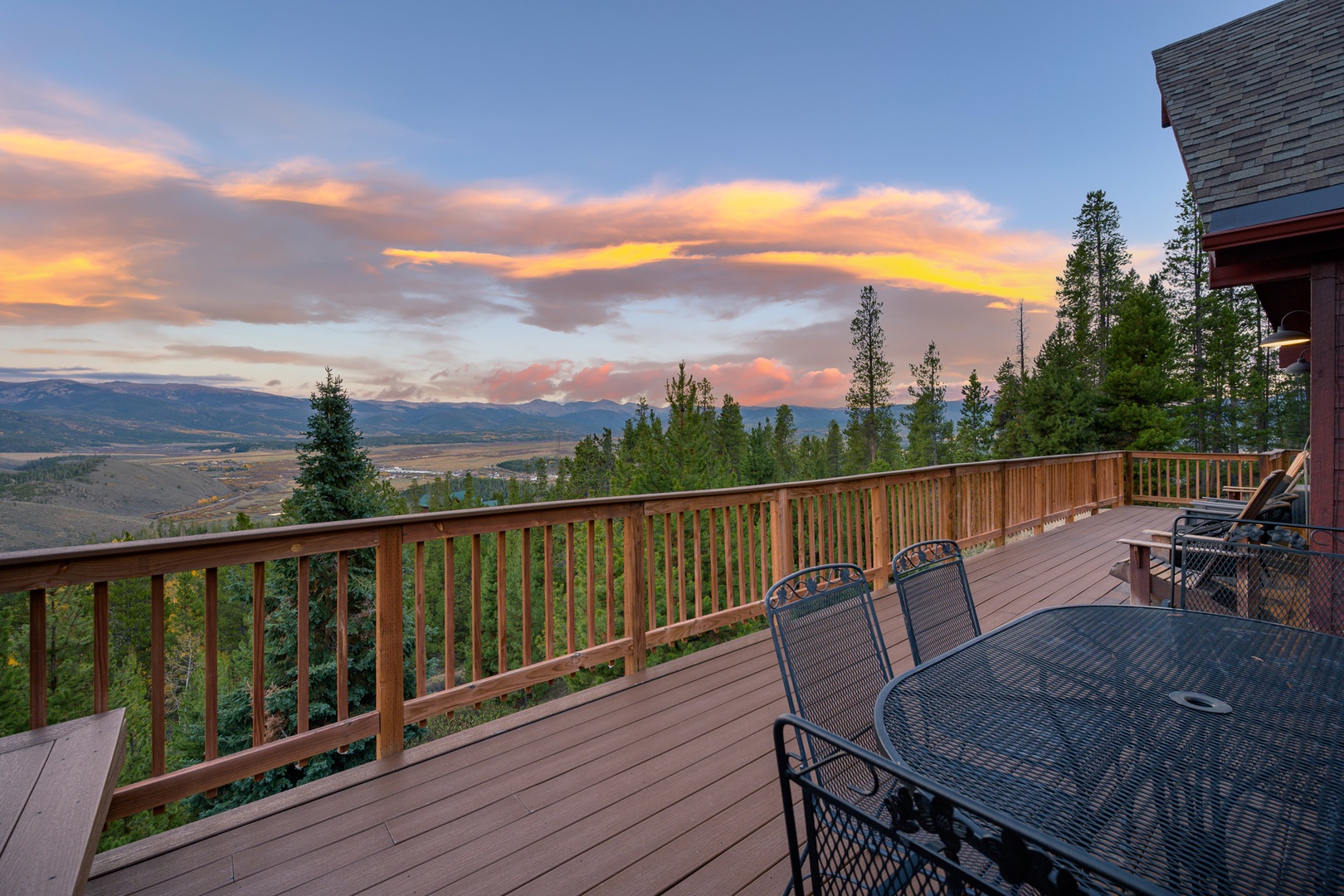 A spacious deck with a dining table and lounge chairs overlooks a breathtaking mountain valley as the sky glows with golden sunset hues. Surrounded by tall pines and open views, this spot invites you to unwind and soak in the natural beauty.