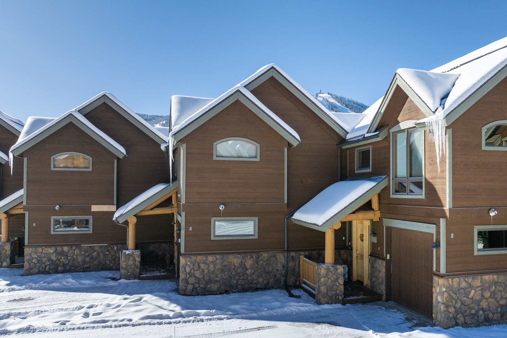Snow-dusted mountain chalets feature stone accents and covered entryways, set against a backdrop of forested peaks under clear blue skies.