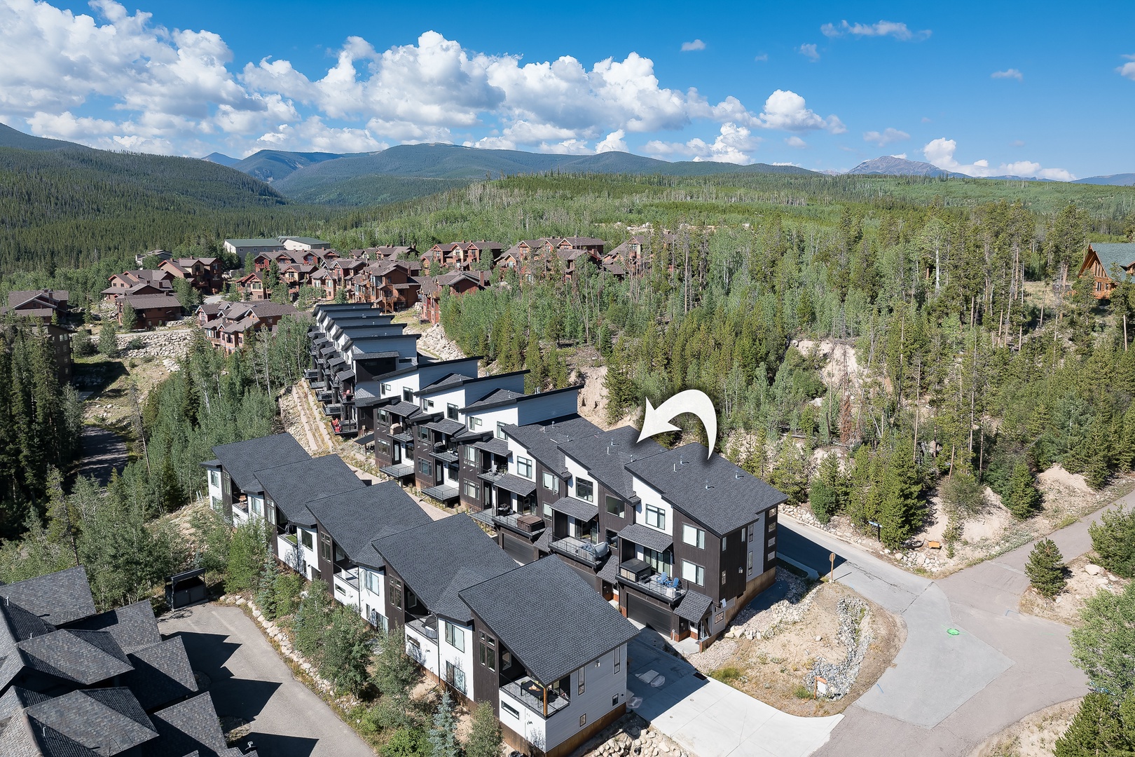 Aerial view of modern mountain townhomes nestled within a forested alpine community, surrounded by dense evergreens and dramatic mountain peaks.