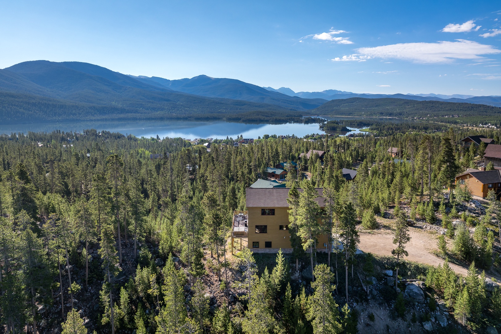 Aerial view of the property nestled among tall pines, with a serene lake and majestic mountain peaks visible in the distance—a peaceful natural setting.