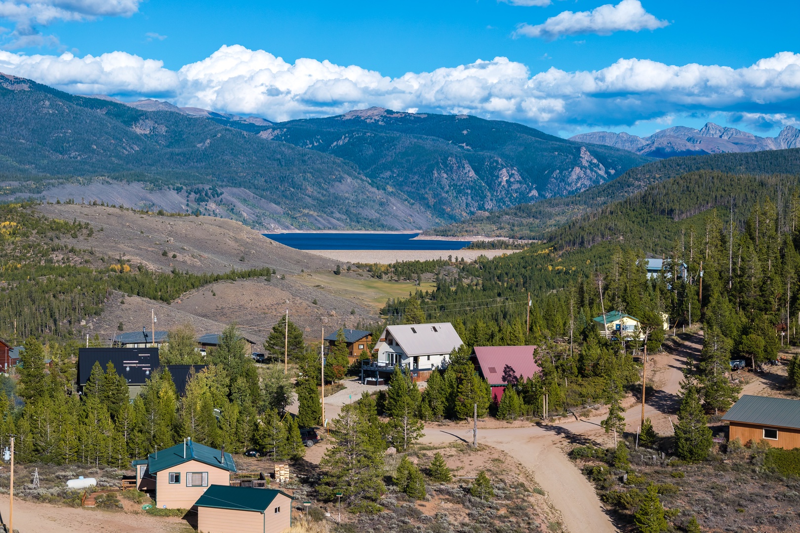 An aerial view shows the property nestled among tall pines in a mountain valley, with a pristine lake and forested peaks visible in the distance—a peaceful setting close to nature.