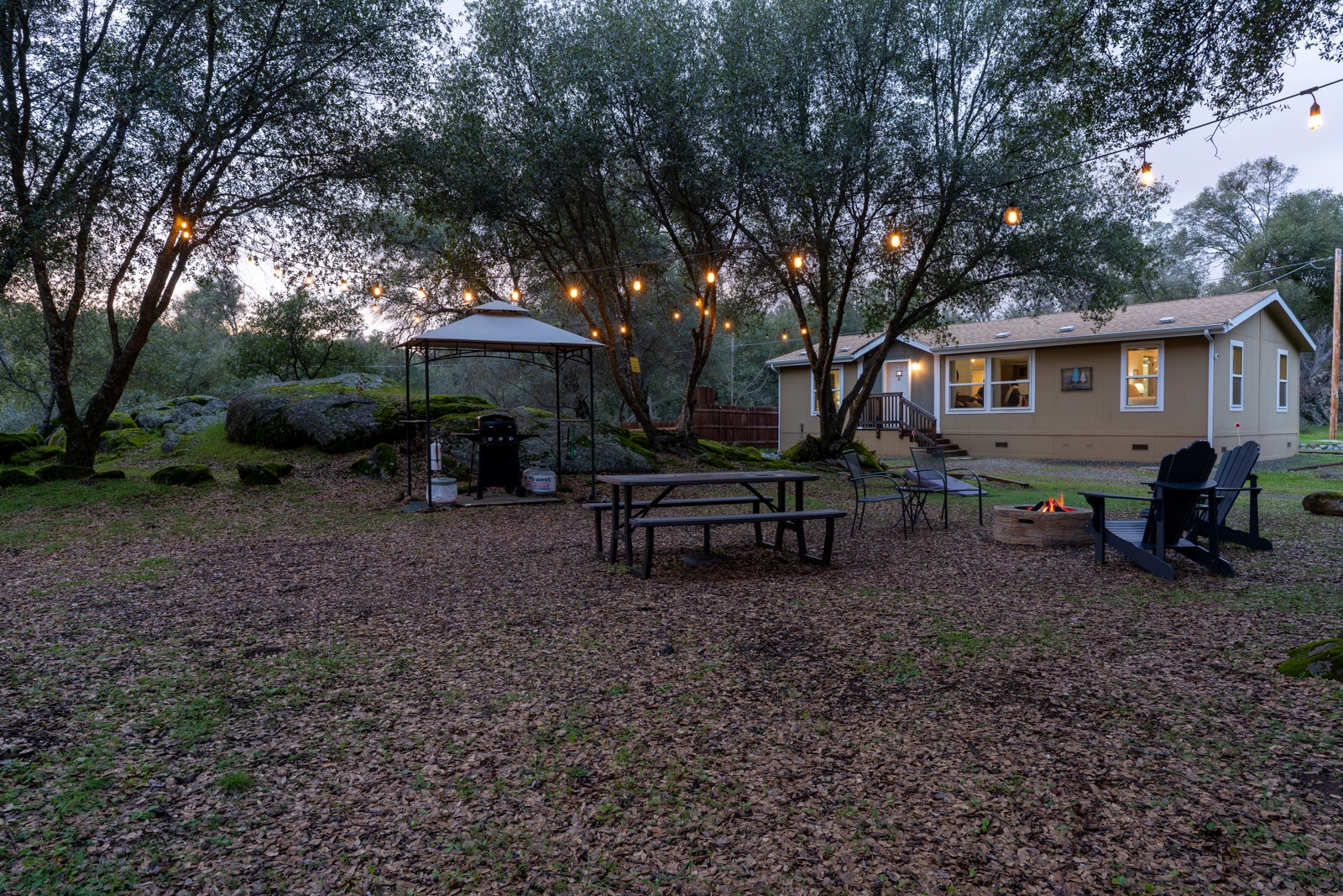 Cozy property nestled among trees with outdoor dining, BBQ area, and fire pit for memorable evening gatherings under twinkling string lights.