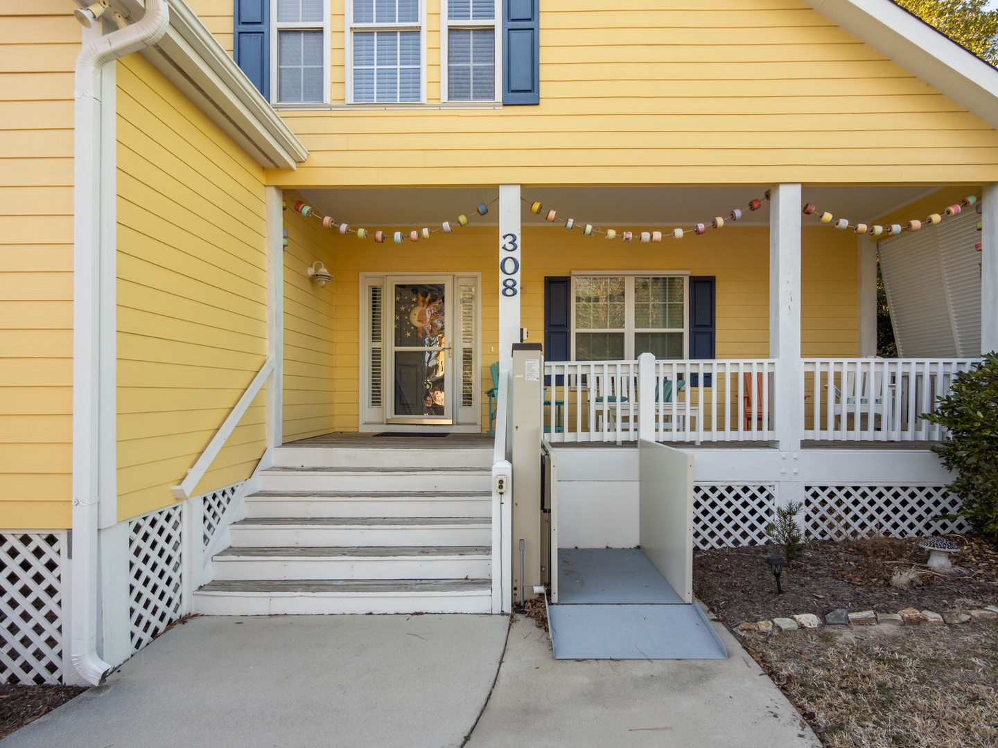 Charming yellow cottage with cheerful porch featuring festive string lights and accessible ramp entrance.