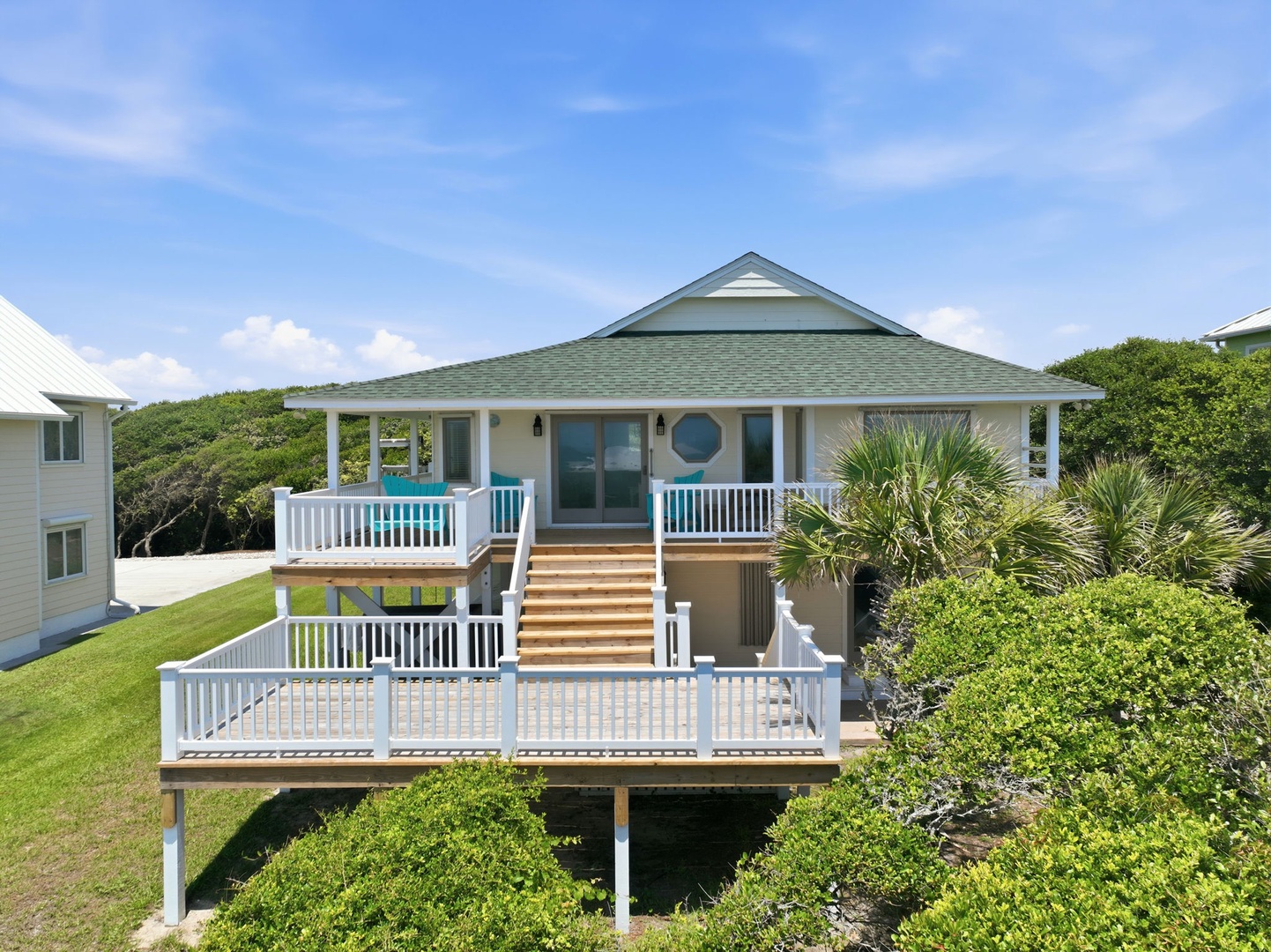 A coastal cottage on stilts features a covered wraparound porch, multiple deck levels, and lush tropical plantings set against blue skies and neighboring beach homes.