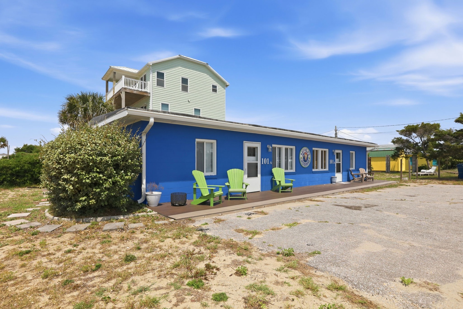 The vibrant blue building and neighboring green house create a colorful coastal arrival point, with numbered doors opening to your beachside escape and cheerful Adirondack chairs promising relaxation.