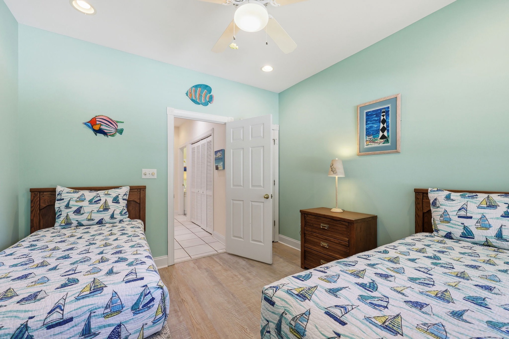 Drift off to sleep in this cheerful coastal-themed bedroom with two cozy twin beds, playful sailboat linens, and whimsical fish art creating a serene seaside escape.