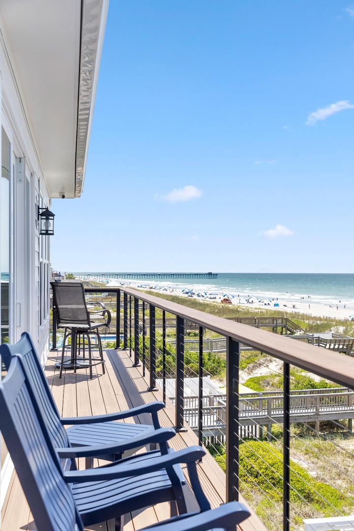 Expansive beachfront balcony offers panoramic views of sand, surf, and horizon, with Adirondack chairs and bar seating for watching waves and sunsets in comfort.