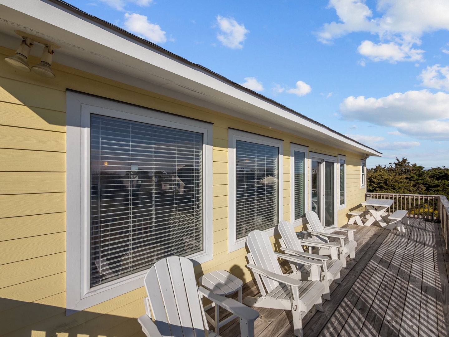 Cheerful yellow coastal cottage exterior featuring a spacious deck with classic Adirondack chairs overlooking surrounding greenery under bright blue skies.
