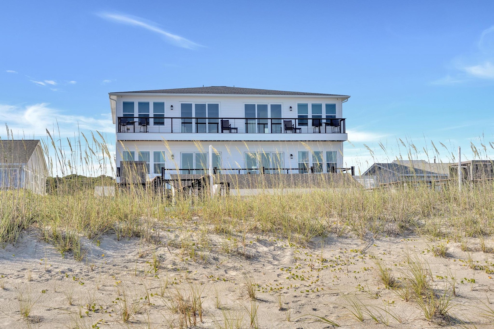 Beachfront property featuring direct sandy beach access with dunes and beach grasses leading to a multi-level coastal building with private balconies under clear blue skies.