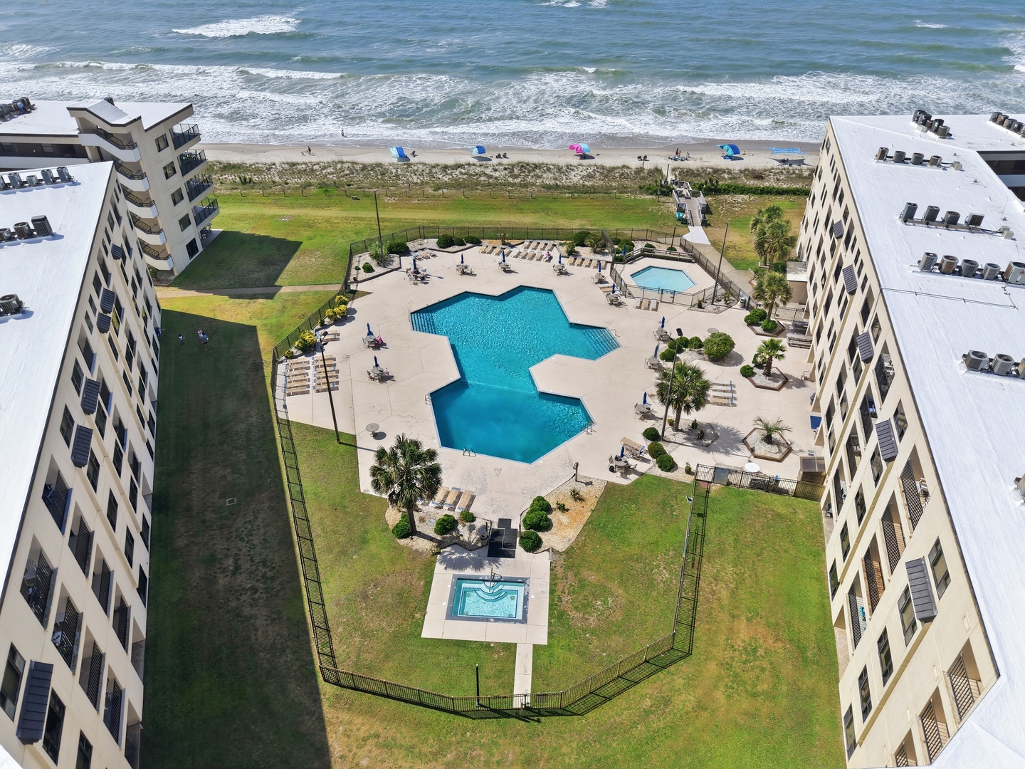 Aerial view of the beachfront property showcasing dual swimming pools, a hot tub, and lounge area nestled between residential buildings with direct beach access and ocean beyond.