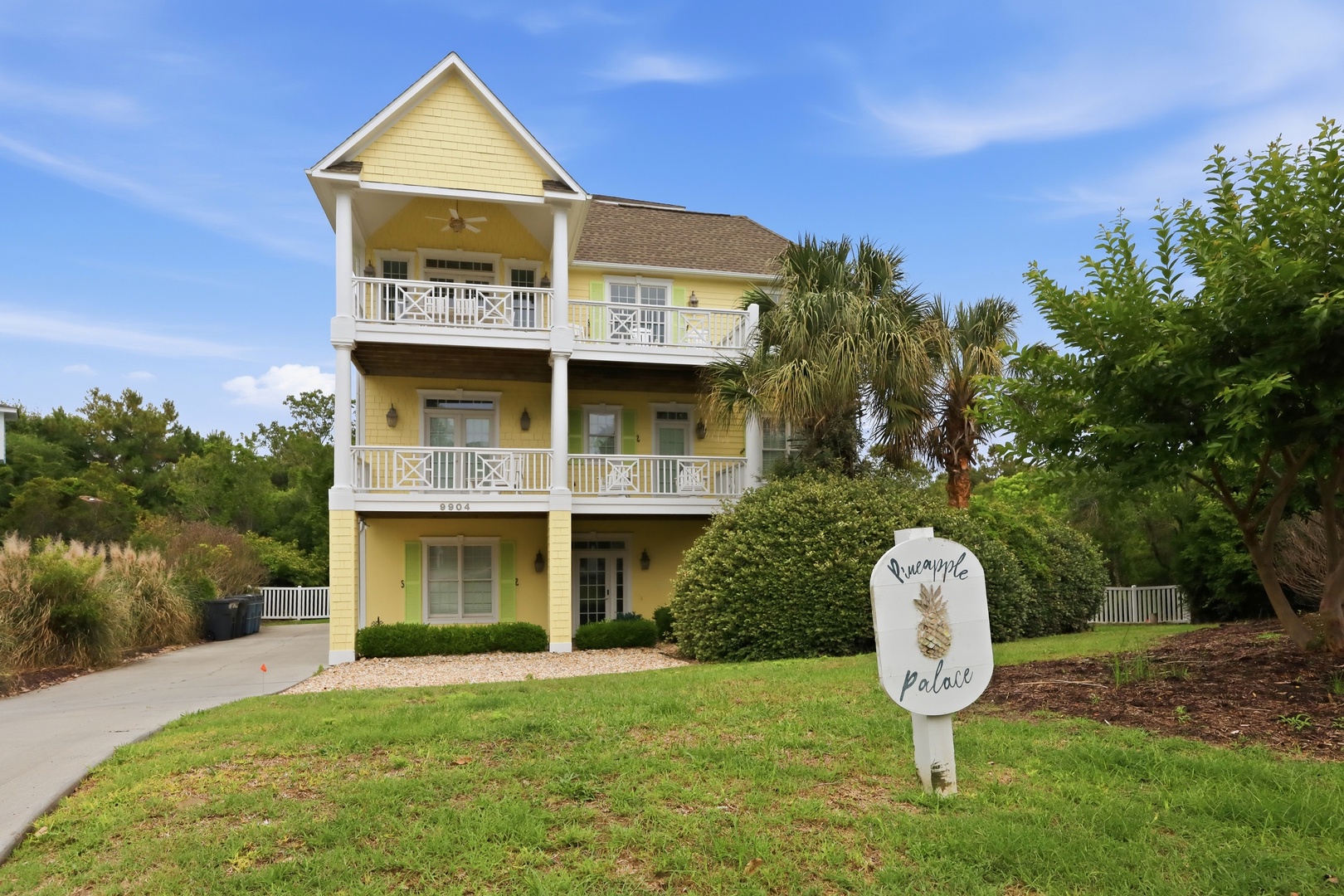 Pineapple Palace welcomes guests with cheerful yellow siding, multiple balconies, and lush coastal landscaping under clear blue skies—a bright, inviting arrival experience.