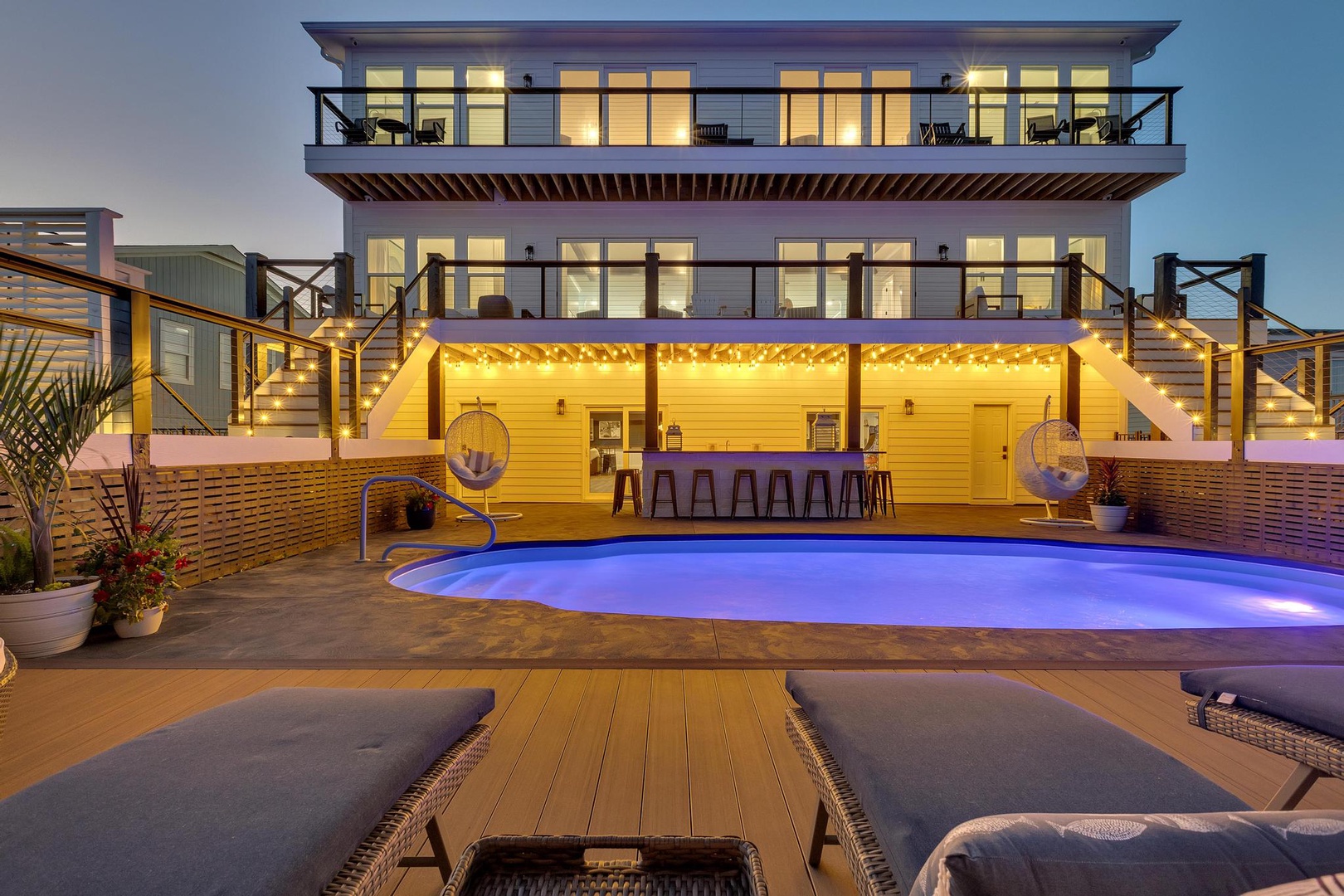 The modern three-story property features expansive balconies on each level, an illuminated pool with integrated bar seating, and an inviting outdoor entertainment area perfect for sunset gatherings.