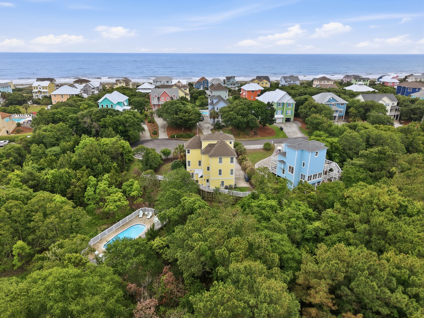 Aerial view of a coastal vacation rental community featuring colorful beach homes nestled among lush green trees, with ocean views and a private pool on-site.