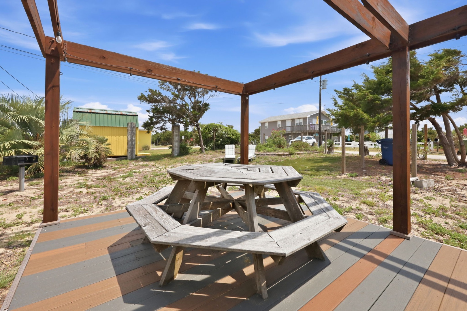Outdoor communal picnic area with pergola shelter and hexagonal table, located in a coastal residential neighborhood with palm trees and nearby buildings.