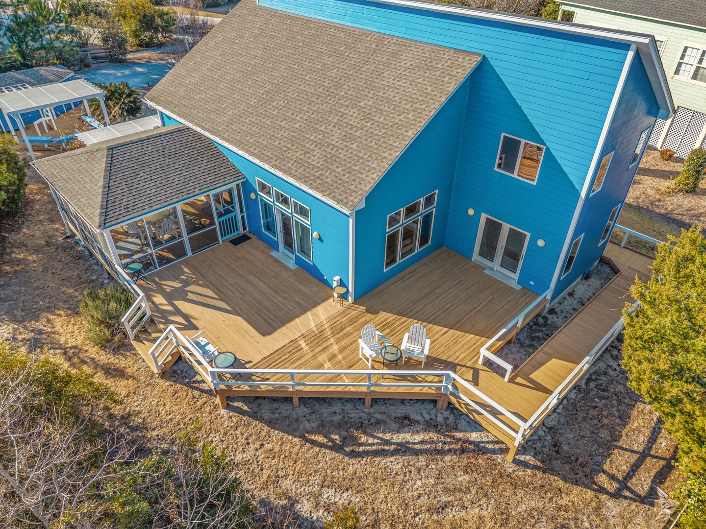 Modern blue beach house featuring expansive wraparound deck with Adirondack chairs, perfect for morning coffee and sunset views in peaceful residential setting.