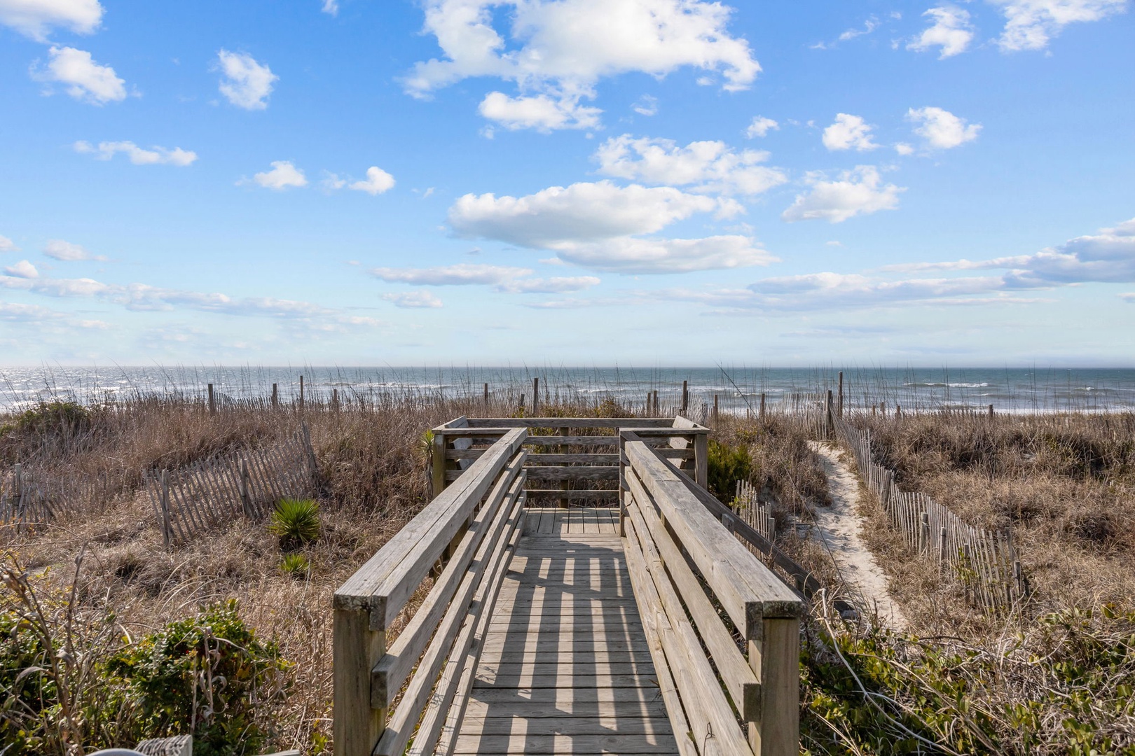 A wooden boardwalk leads through coastal dunes to the beach, with ocean views and blue skies creating a perfect pathway to relaxation.