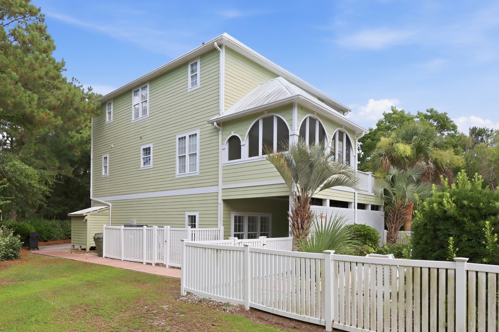 A charming three-story coastal home with soft green siding and white trim, featuring covered parking and a white picket fence, surrounded by lush palms and mature trees in a peaceful neighborhood setting.