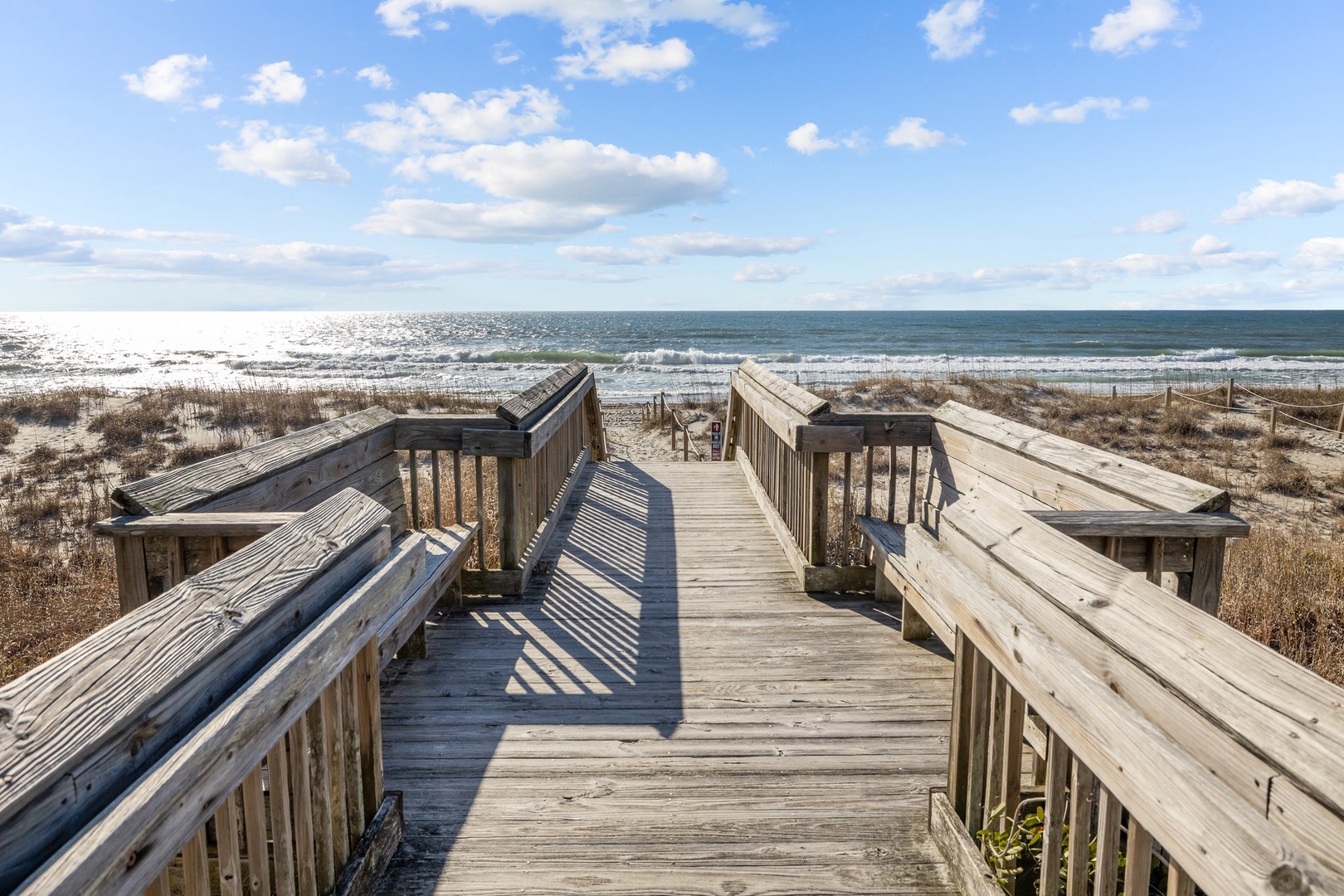 Wooden boardwalk leads directly to pristine sandy beach and sparkling ocean waves under a beautiful blue sky.