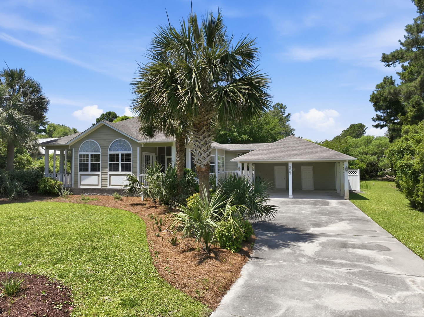 A charming coastal-style home sits on a lush lot, framed by mature palms and tropical landscaping. Spacious two-car garage and inviting front porch welcome guests to a peaceful retreat.