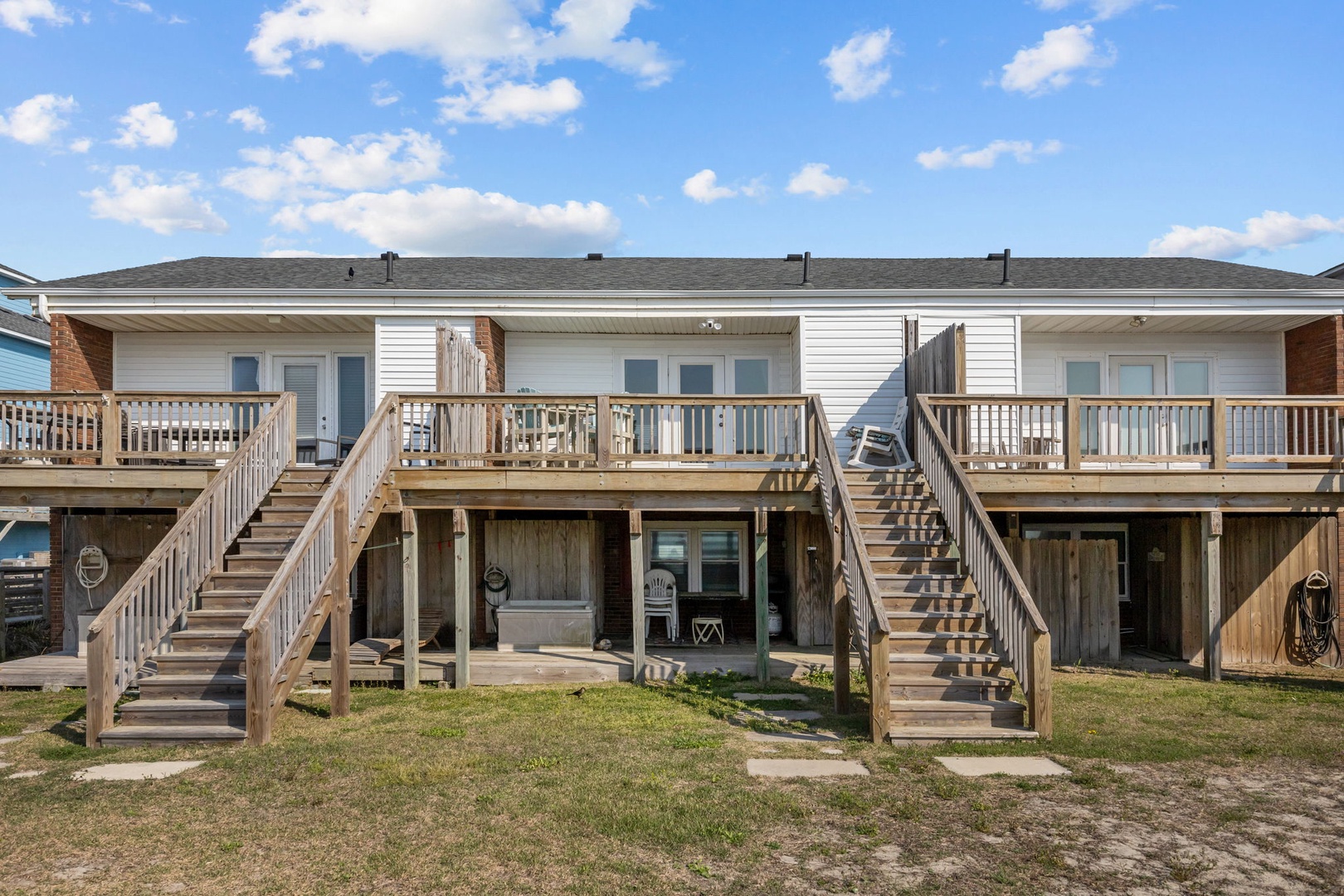 A beachside duplex with dual elevated decks and ground-level patios, surrounded by coastal grasses under bright blue skies.
