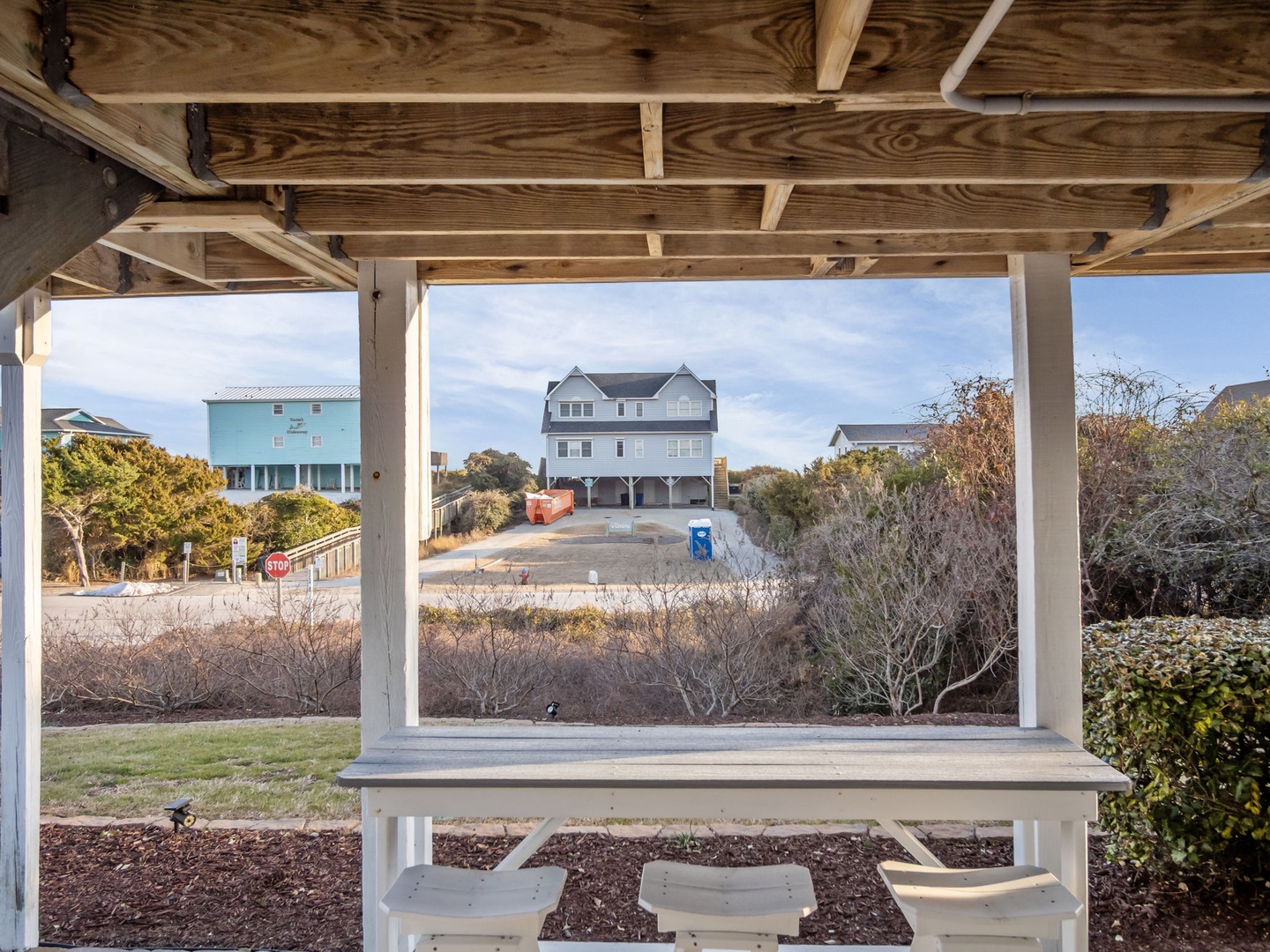 Covered outdoor dining area with wooden beams and white table, surrounded by coastal homes and natural vegetation.
