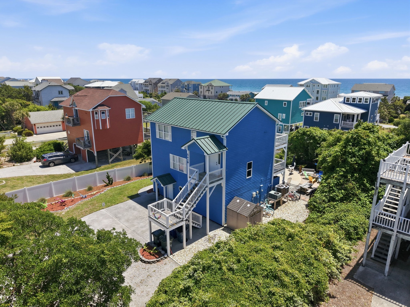 Aerial view of a bright blue coastal rental home featuring elevated construction, covered parking, outdoor patio space, and a quiet beachside neighborhood setting with ocean views in the distance.