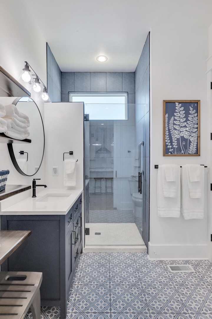 Your private bathroom blends modern elegance with spa-like serenity. Crisp white cabinetry, patterned tile floors, soft towels, and a sleek glass-enclosed shower invite you to unwind.