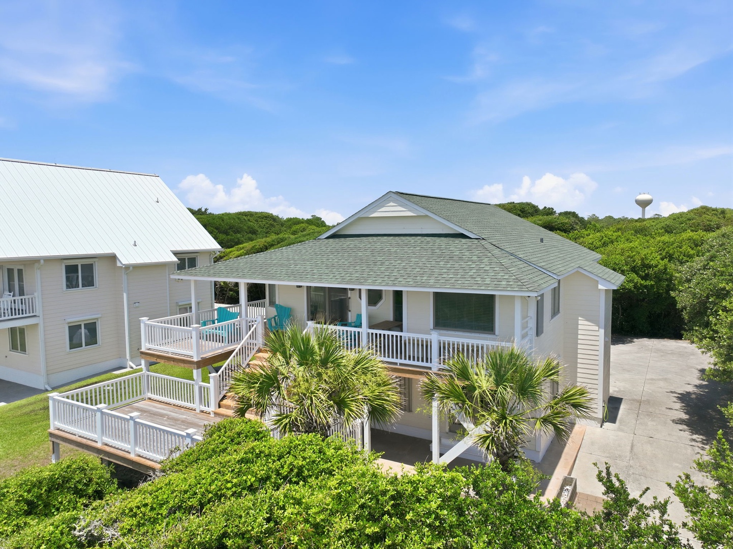 Elevated beach cottage with spacious wraparound decks and coastal landscaping, situated among lush greenery near the shore.