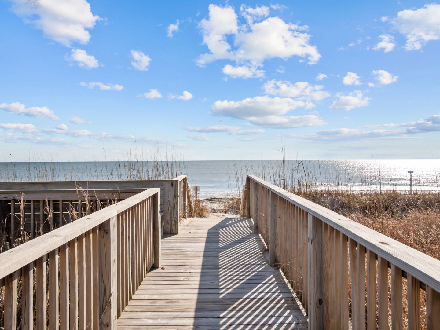 Wooden boardwalk leads directly to pristine sandy beach and sparkling ocean waters under bright blue skies.
