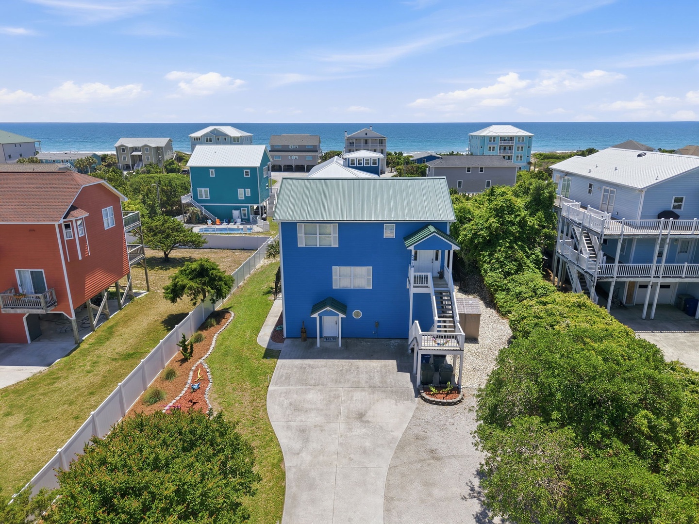 Aerial view of a blue coastal property situated in a quiet neighborhood, just steps from the ocean, surrounded by lush greenery and nearby vacation homes.