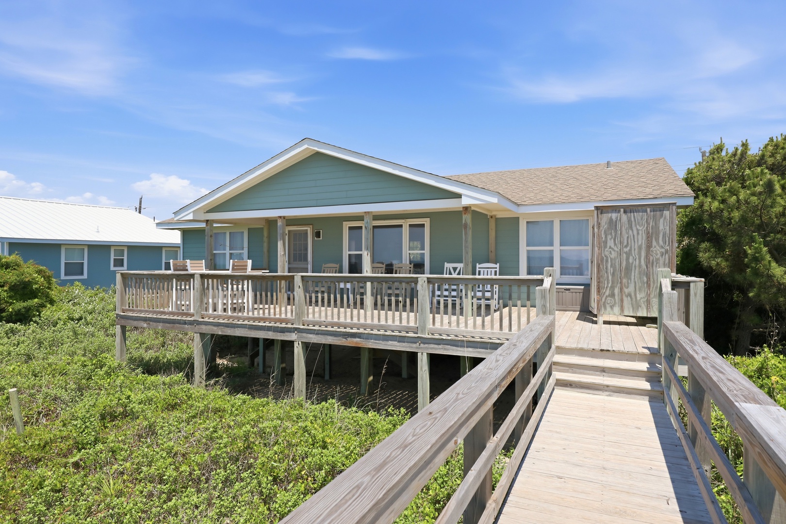 Charming coastal beach house with an elevated deck and private boardwalk access, nestled among natural dunes and greenery under sunny skies.