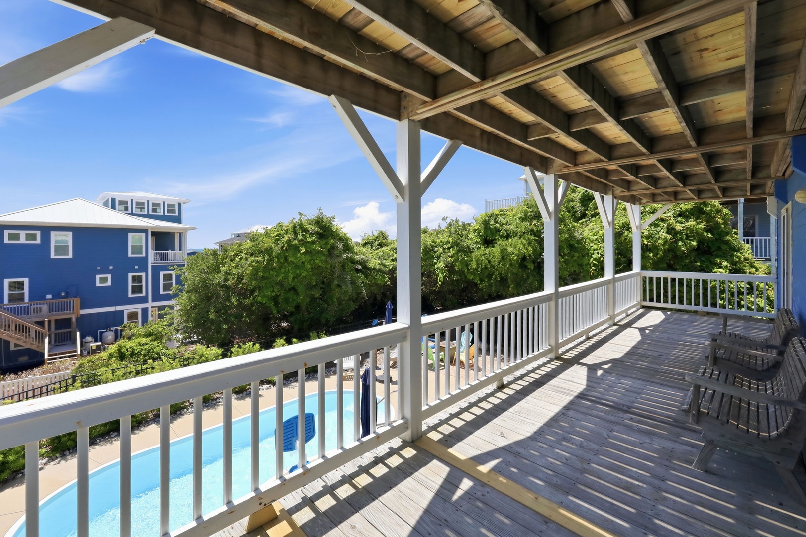 A covered deck featuring poolside views and glimpses of neighboring beach houses, with wicker seating to enjoy this sunny coastal setting.