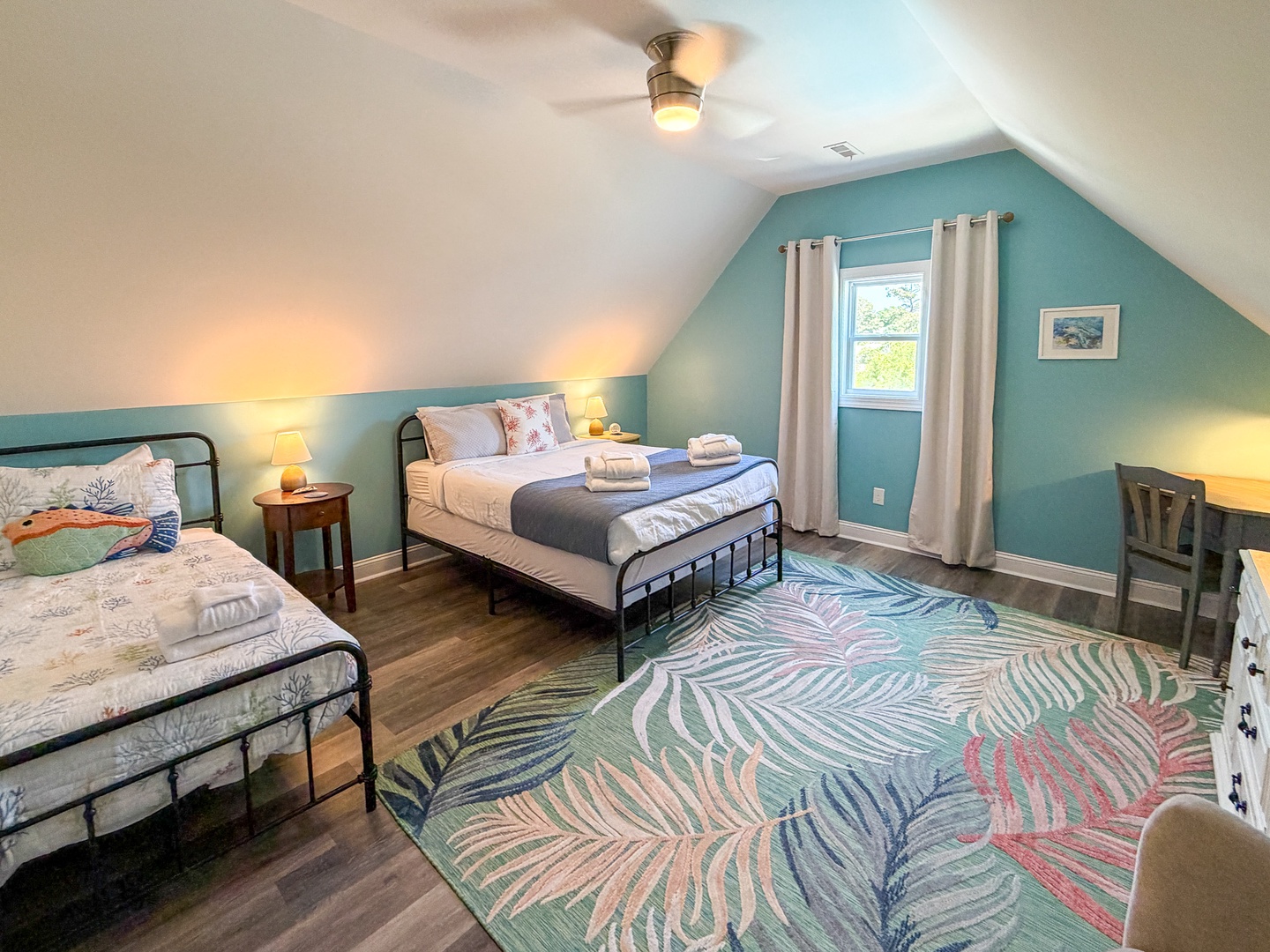Your cozy retreat awaits with two comfortable beds, soothing teal walls, a ceiling fan for comfort, and warm natural light flowing through the window. A peaceful space to recharge and relax.