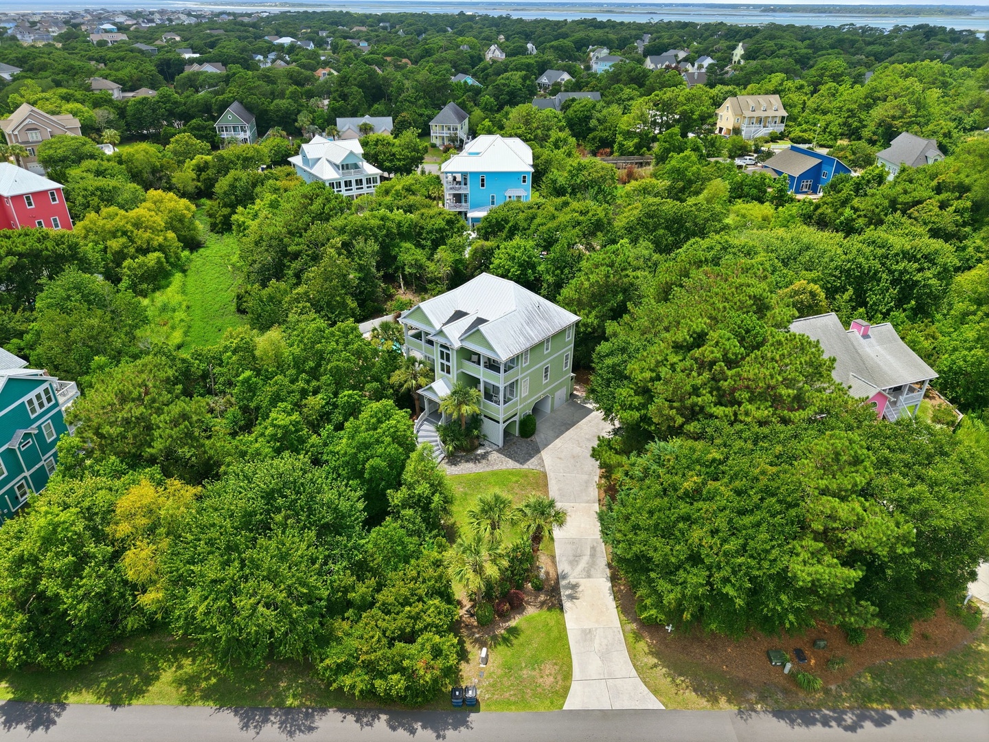 Aerial view of a charming coastal neighborhood featuring a green cottage surrounded by lush summer foliage, with glimpses of waterfront beyond.
