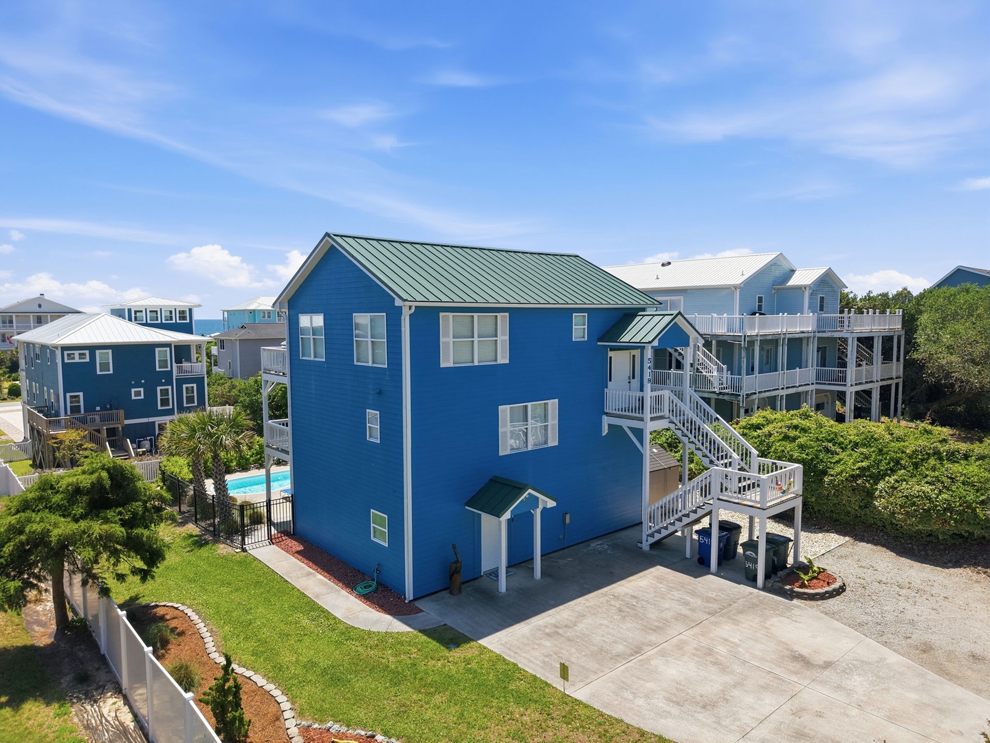 Bright blue coastal home elevated on stilts with white railings, private parking, and lush tropical landscaping in a peaceful beach neighborhood.