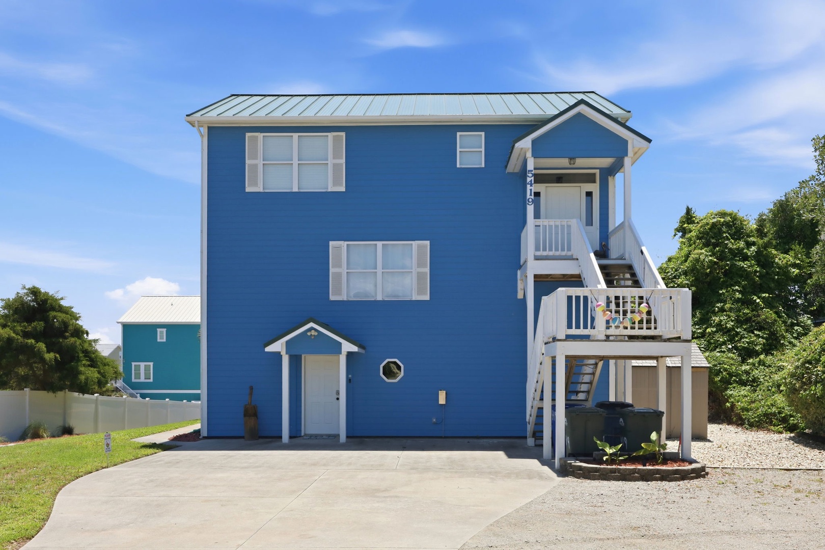 This charming blue coastal home features elevated architecture with private balconies, convenient ground-level parking, and a peaceful residential setting surrounded by natural greenery.